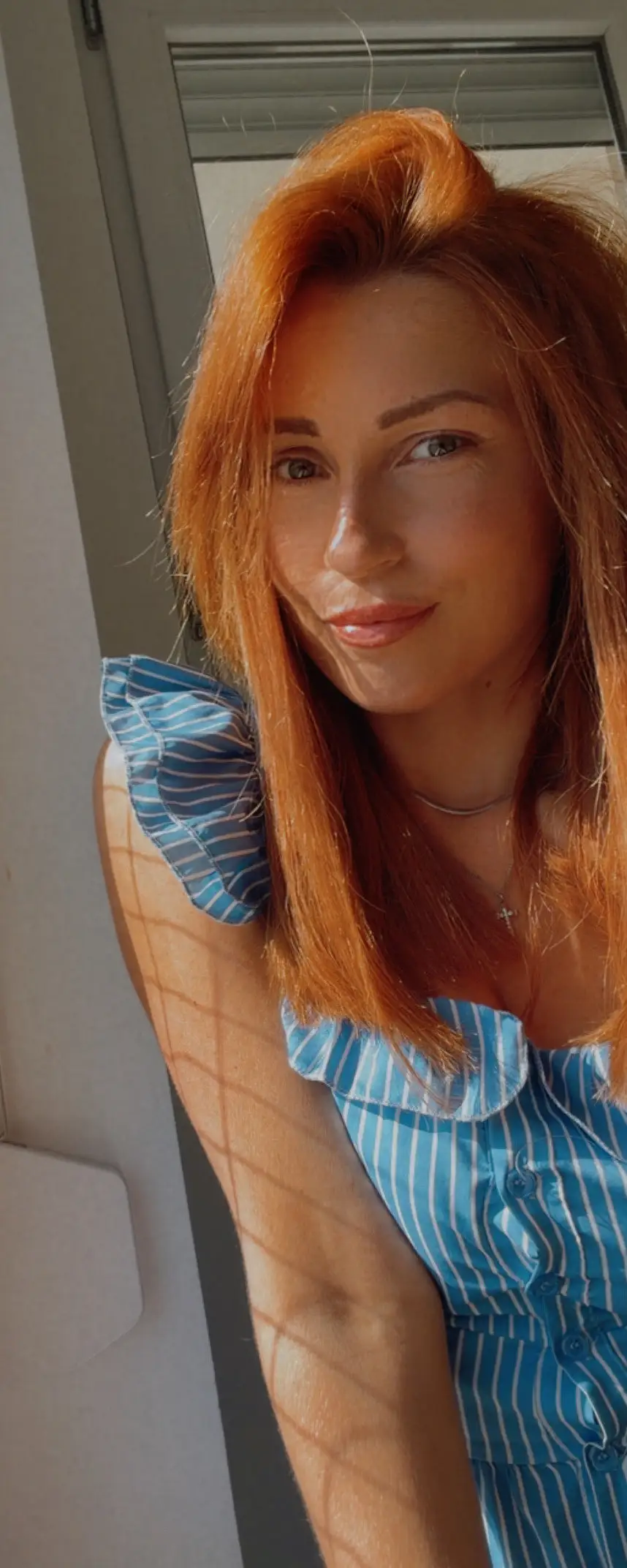Milica leaning in a sunlit doorway, with patterned shadow falling across her arm and the wall beside her.